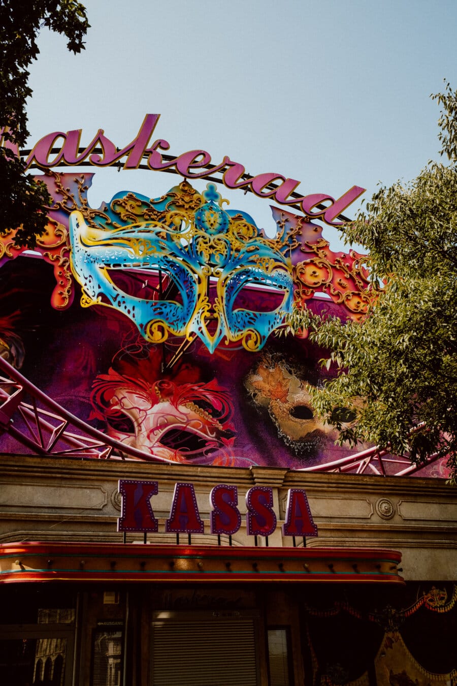 A bustling carnival booth under a clear blue sky, with a large, colorful masquerade mask decoration. The word "Kassa" (ticket office) is clearly visible under the mask. The booth is surrounded by greenery, adding to the festive atmosphere.  