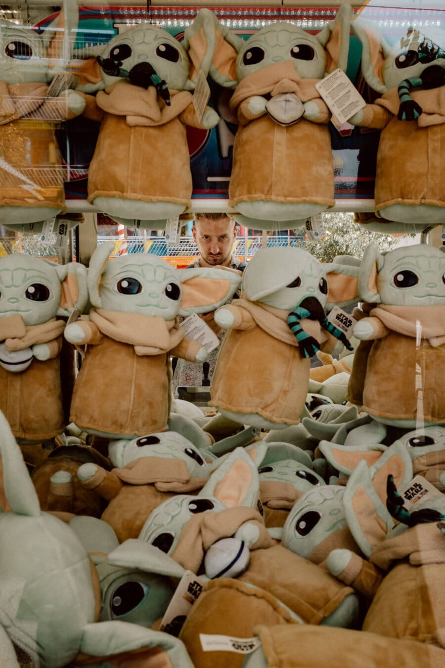 The man is looking at a claw machine filled with numerous plush toys resembling a small Yoda. The plush toys have large eyes, green skin and wear brown robes with a brown and turquoise scarf. The man seems focused as he looks over the assortment of toys.  