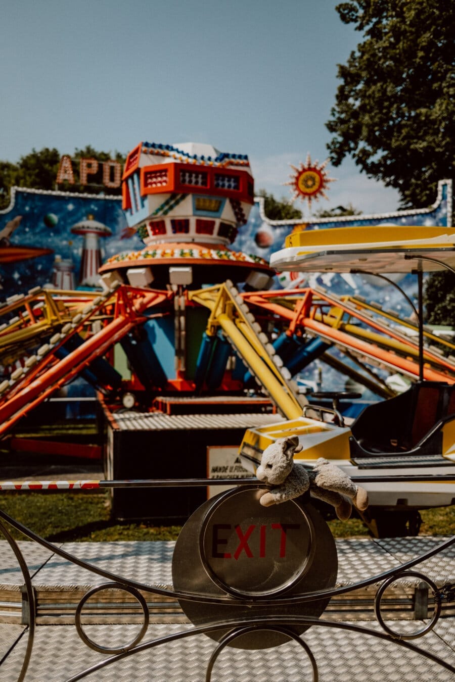 A colorful amusement park ride called "Apollo" is in the background against a clear blue sky. In the foreground, on a metal railing, is a small stuffed animal with the word "EXIT" below. Trees partially frame the scene.  