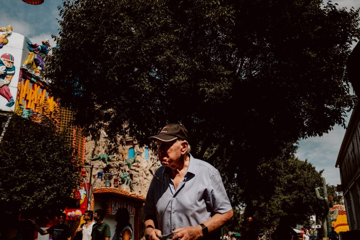 An elderly man in a black cap and striped shirt stands outside, while trees, colorful amusement park rides and amusement park signs can be seen in the background. The sky is partly overcast, and a few people are enjoying the fair. 