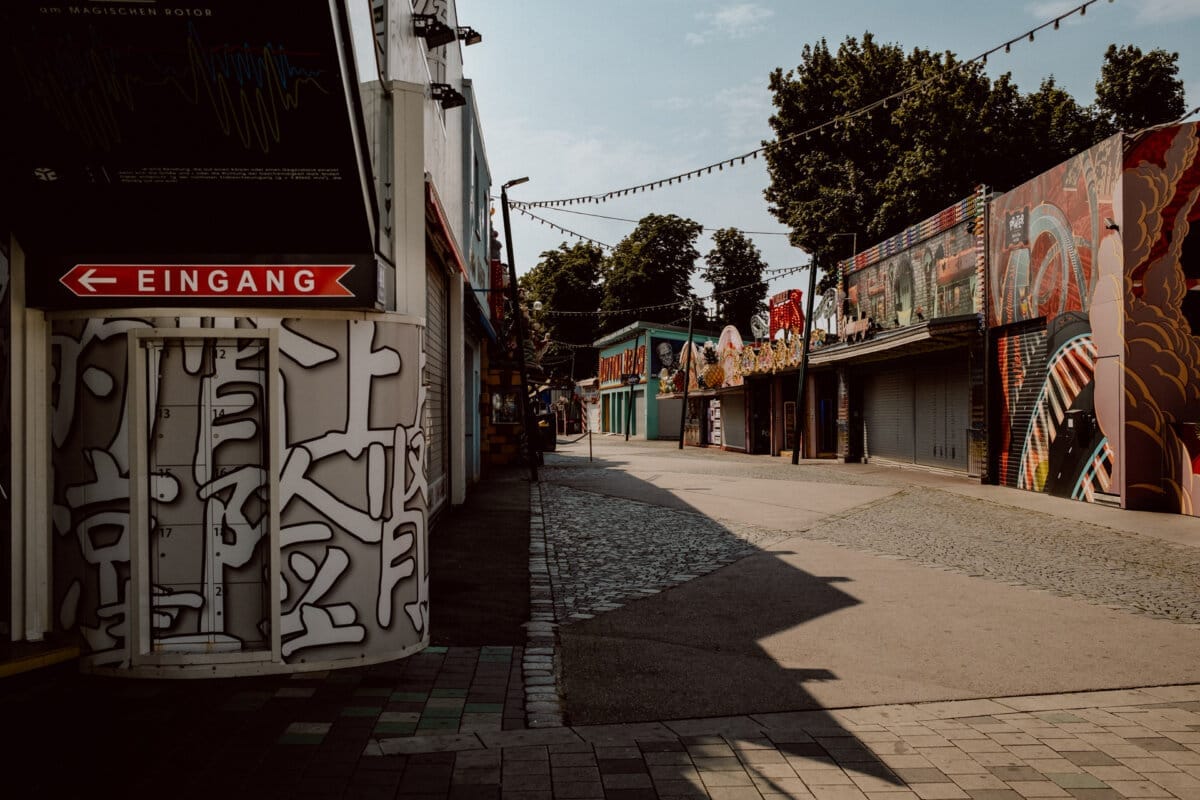 The empty street in the amusement park is dotted with closed, graffiti-covered stores and colorful murals. A sign reading "EINGANG" (German for "entrance") points to the left. The sky is clear and strings of lights hang over the street. Trees can be seen in the background.   