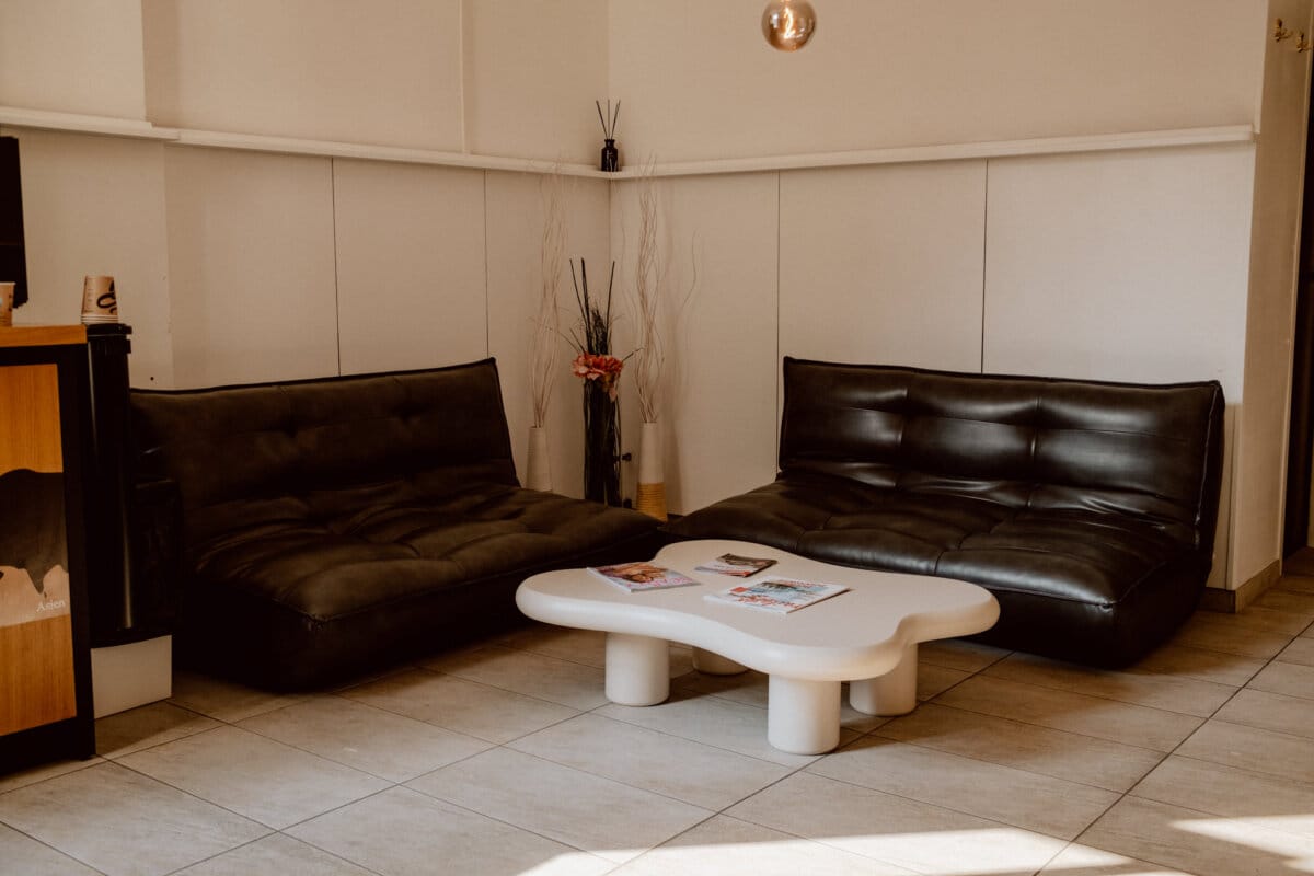 A modern waiting room with two black leather sofas, a white coffee table with an abstract shape in the middle and magazines on the table. The room has beige tiles and minimalist decor, including a vase with tall reeds and a wall-hanging cabinet. 