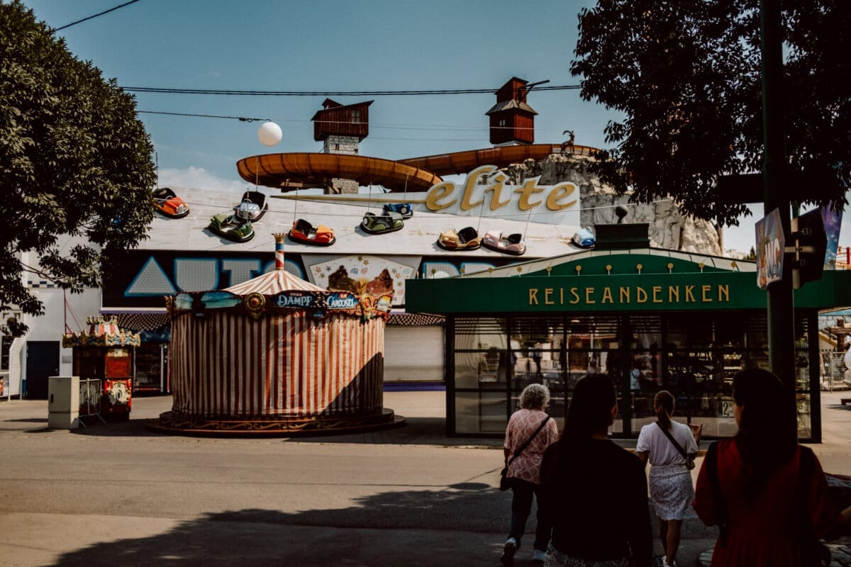 The scene at the attraction fair with a row of colorful bumper cars in front of a building marked "elite." Nearby, a circular tent with vibrant designs stands next to a green store marked "REISEANDENKEN." A few people walk around and watch the attractions.  