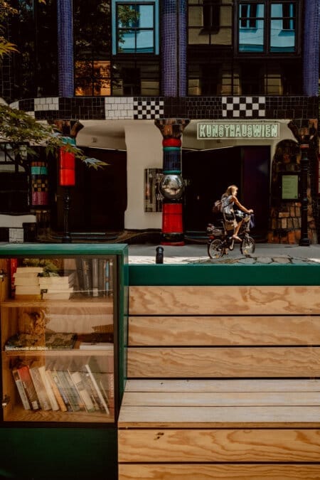 A person rides a bicycle past the entrance to Kunsthaus Wien with its eclectic, colorful facade. In the foreground, a wooden structure with a small public bookshelf filled with various books adds a touch of community spirit to the urban scene. 