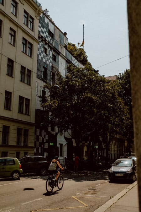 A person rides a bicycle along a street full of parked cars, and in the background you can see a tall, uniquely decorated building with a patchwork pattern of black, white and gray tiles. Trees partially obscure the building, and several people are walking nearby. 