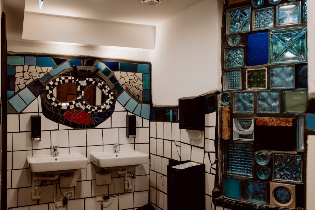 A bathroom with two white sinks and mirrors, surrounded by colorful mosaic tiles on the walls. The mosaic includes various shades of blue, green and other colors, creating an artistic and lively atmosphere. 