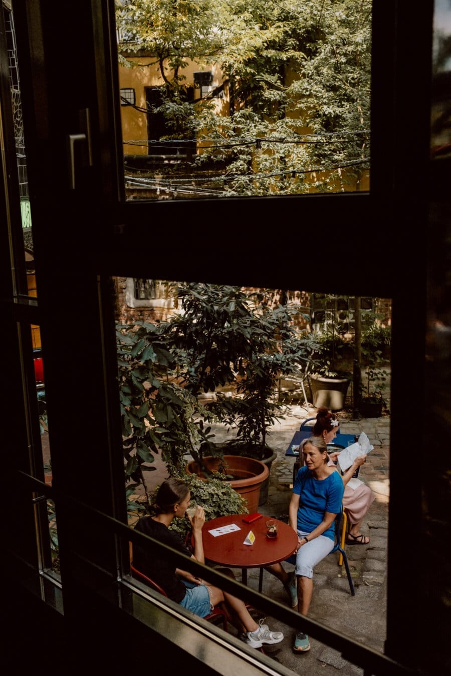 The view from the window is of four people sitting at a red table in the garden courtyard. They are surrounded by greenery and potted plants, enjoying the outdoor conversation. The scene is peaceful and shaded by nearby trees.  