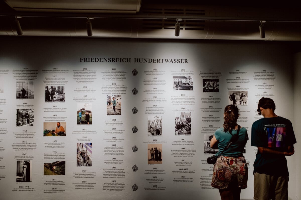 Two people stand in front of the museum's wall exhibition, which details the life and work of Friedensreich Hundertwasser. The exhibit includes photographs, artwork and key dates from the 1920s to 2000. Visitors are engaged in reading the information.   