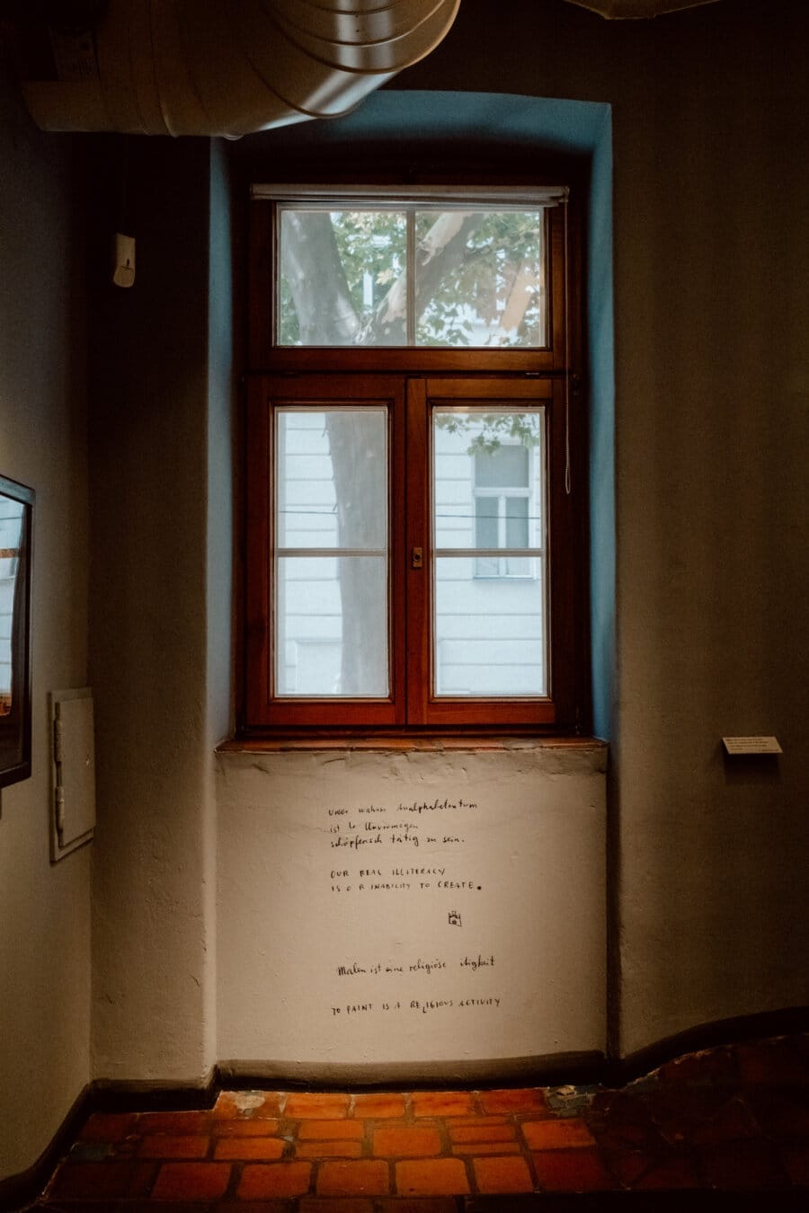 The dimly lit corridor has a wood-framed window and a view of the wall and windows of a distant building. Below the window, the written text on the wall is visible. The floor is covered with reddish-brown tiles, and part of a picture frame can be seen to the left.  