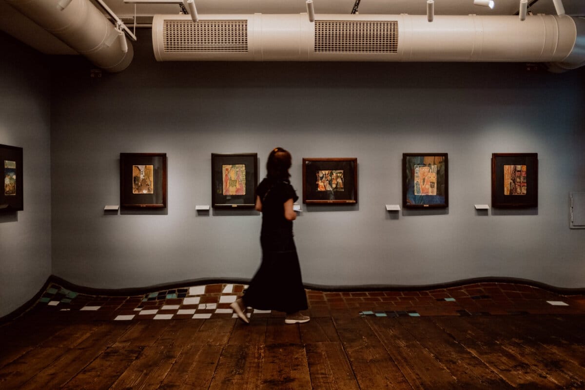 A person in a long, dark dress walks through an art gallery with wooden floors, looking at framed works of art displayed on a blue-gray wall. The gallery has bright lighting with visible air ducts on the ceiling. 