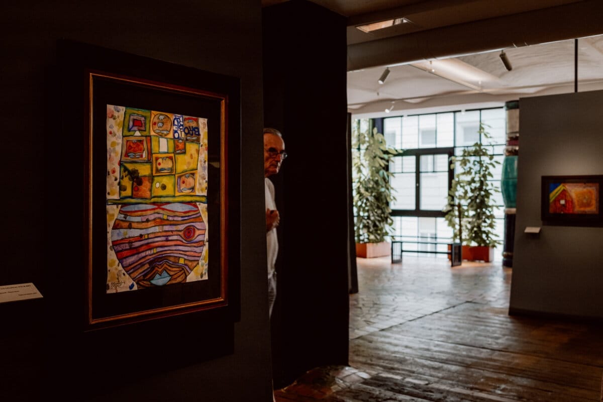 A man looks out from behind a partition wall in an art gallery. In the foreground is a brightly colored abstract painting, and the background opens up to a spacious, well-lit room with windows and potted plants. Another work of art is visible on the far wall.  