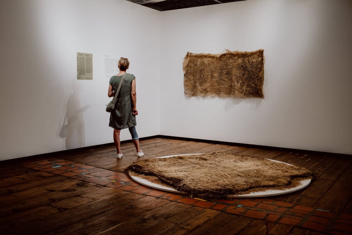 A person with short hair observes an exhibit in an art gallery. She is wearing a sleeveless dress and sneakers. The exhibits include a rectangular textured wall hanging and a large round textured piece lying on the floor. The room has wooden floors and white walls.   