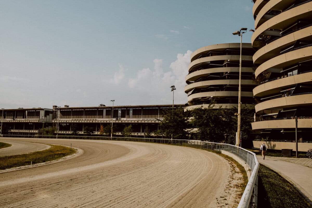 The curved dirt racetrack is surrounded by metal barriers, leading to modern, multi-story circular parking lots on the right. The sky is clear, with a few clouds. A single person walks near the base of the building, adding a sense of scale to the sprawling scene.  