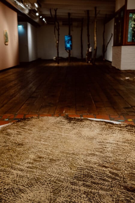 A dimly lit room with a wooden floor and vertical wooden pillars distributed unevenly throughout the space. In the foreground is a round, textured carpet, while a large blue screen is visible on the opposite wall. The walls and ceiling beams appear unfinished.  