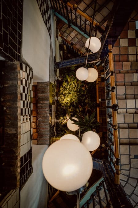 A view down a spiral staircase lined with blue and black tiles and wooden handrails. Large spherical pendant lamps hang in the middle, illuminating the greenery below. The tiles showcase a variety of colors and patterns, creating a vibrant mosaic effect.  