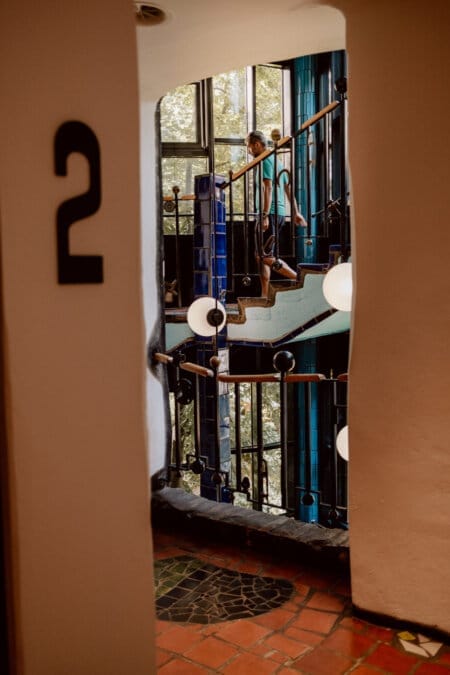 A person ascends a colorful, winding staircase with unique architectural elements, including circular lights and curved handrails. The scene is viewed from a door marked "2," and the background depicts large windows overlooking the greenery outside. 