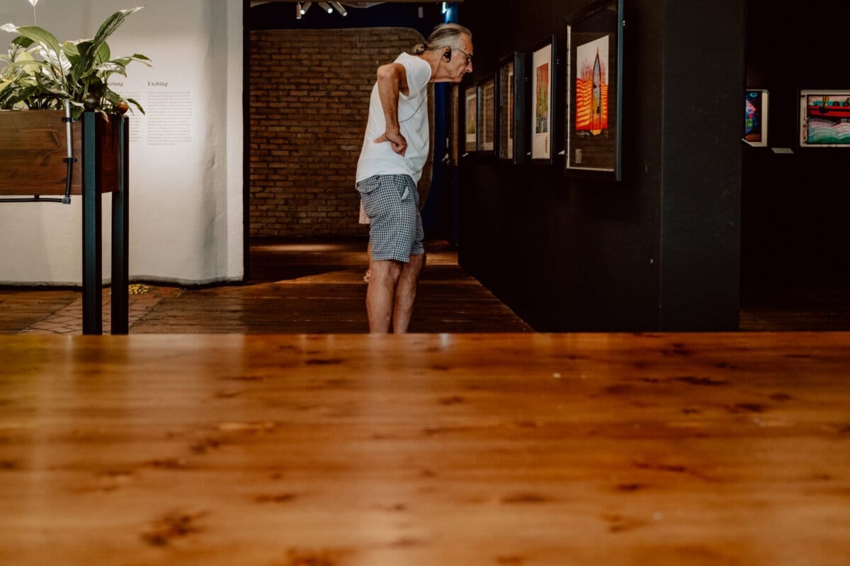 A man with long hair tied up in a ponytail, wearing a white T-shirt and striped shorts, leans over to look at a work of art on a wall in a dimly lit gallery. A plant is visible on the left, and a wooden surface is in the foreground. 