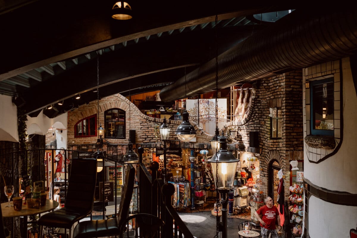 A cozy indoor market with exposed brick walls that houses various stores and restaurants. There are hanging lights, tables with chairs and a person in a red shirt sitting at a table. The view from above shows a second level with additional seating and stores.  