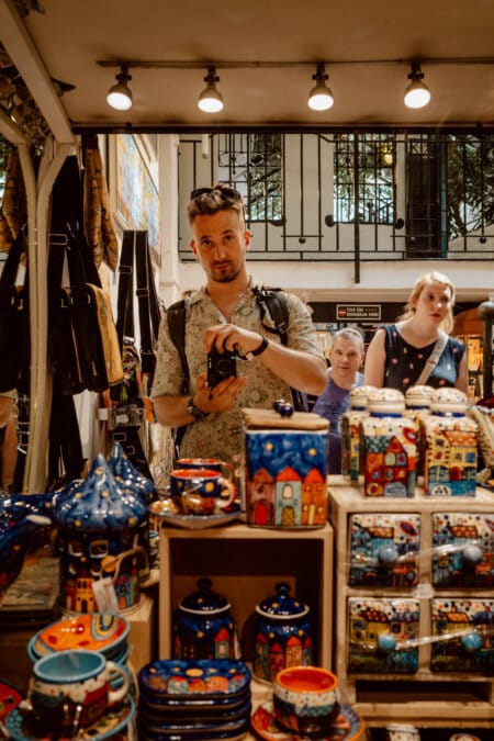 A person wearing a bright patterned shirt takes a photo in a store full of colorful ceramic objects, including vases and mugs, decorated with house designs. In the background, two people are looking at the items on display. The store has bright overhead lighting.  