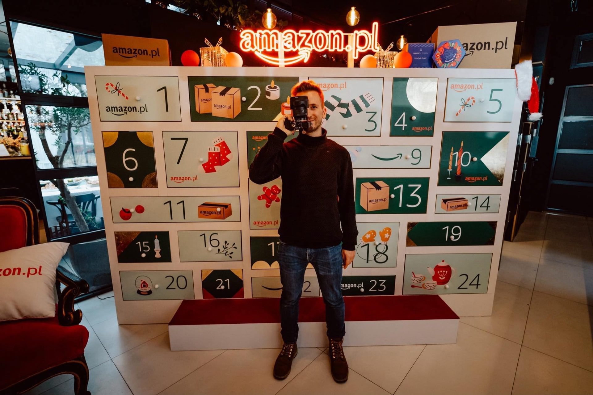 A person stands in front of a large advent calendar display featuring the Amazon.pl logo, captured by event photographer Marcin Krokowski. The calendar has numbered doors and festive decorations, with a neon sign above reading "amazon.pl." The setting appears to be indoors.