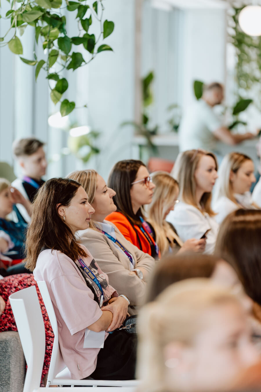 Grupa ludzi siedzi w rzędach, uważnie słuchając prezentacji lub wykładu. W pomieszczeniu jest jasno, z naturalnym światłem, a nad głowami wisi zieleń. Niektórzy noszą smycze z identyfikatorami.