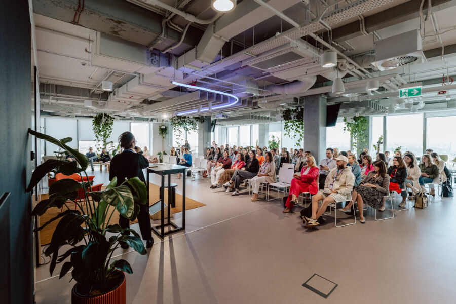 A person speaks at a podium to a seated audience in a modern, open-plan office space. The room has large windows, plants and exposed ceilings with industrial lighting. The audience appears to be engaged in the presentation.