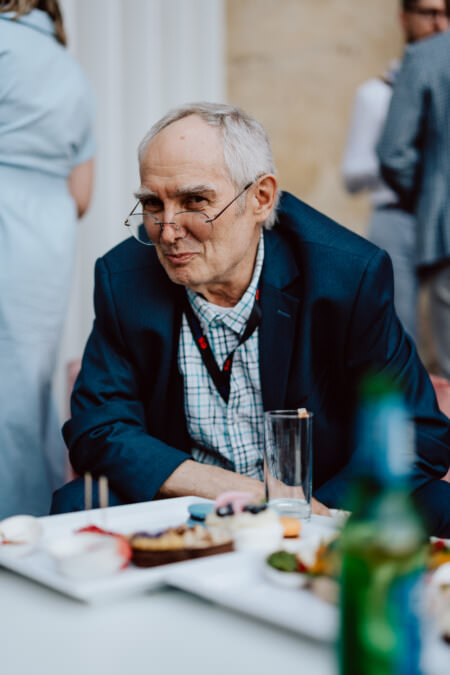 Ein älterer Mann mit Brille und blauem Blazer sitzt an einem Tisch mit einem halbvollen Glas und Tellern mit Speisen. Er steht in der Mitte, bei einem gesellschaftlichen Ereignis, und lehnt sich leicht nach vorne. Im Hintergrund sind unscharfe Menschen und neutrale Wände zu sehen.