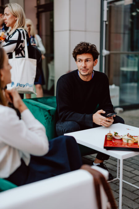 Ein Mann in einem schwarzen Pullover hält ein Telefon in der Hand und sitzt an einem Tisch mit roten Tellern mit Vorspeisen. Er scheint mit einem Gespräch mit einer undeutlichen Person im Vordergrund beschäftigt zu sein. Eine Frau steht im Hintergrund und hält eine PayPal-Tasche in der Hand.