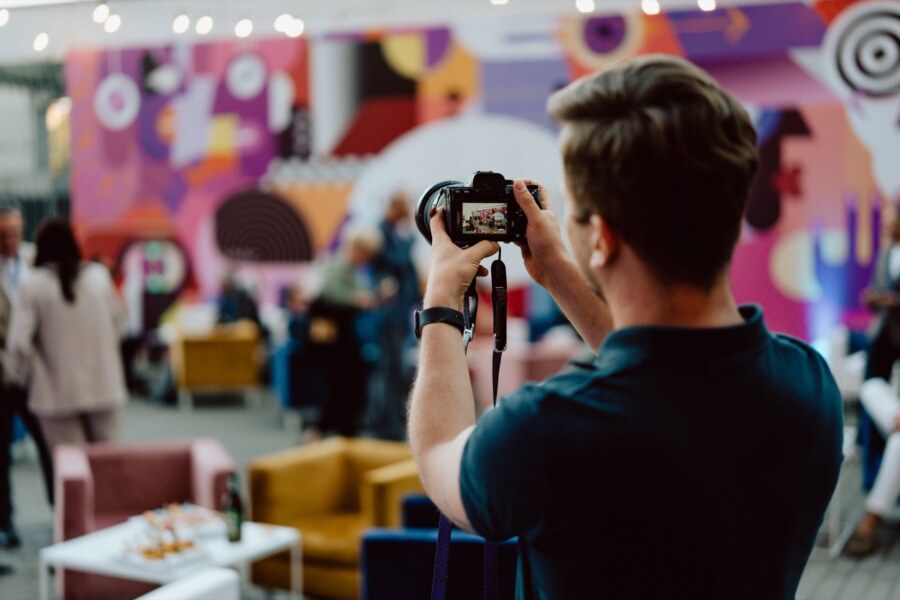 A person takes a picture with a camera at a party. The background is colorful, with abstract art and blurred figures of people mingling. Several chairs and tables are scattered around.