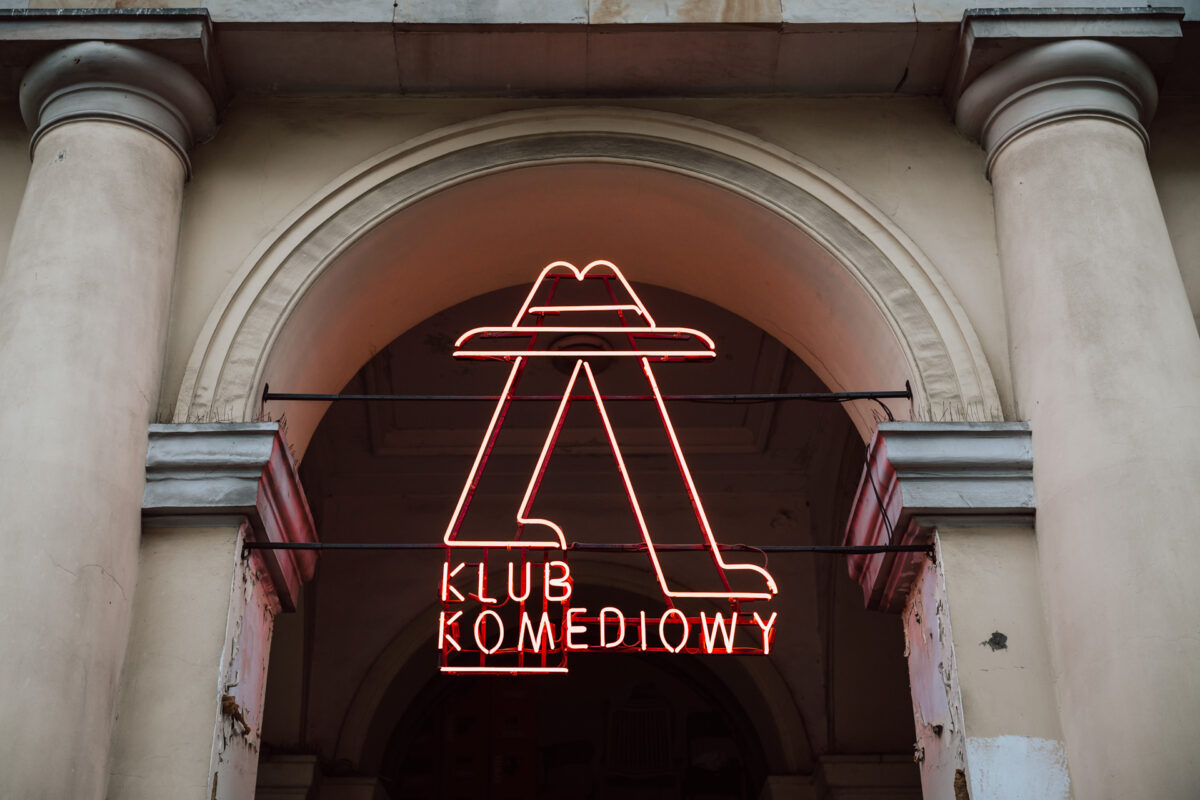 Nie mow do mnie ASAP Klub Komediowy 003 Eine rote Leuchtreklame mit der Aufschrift 'Comedy Club' und dem abstrakten Umriss eines Hutes und Beinen über dem Text ist zwischen zwei Säulen an der beigen Fassade des Gebäudes angebracht.