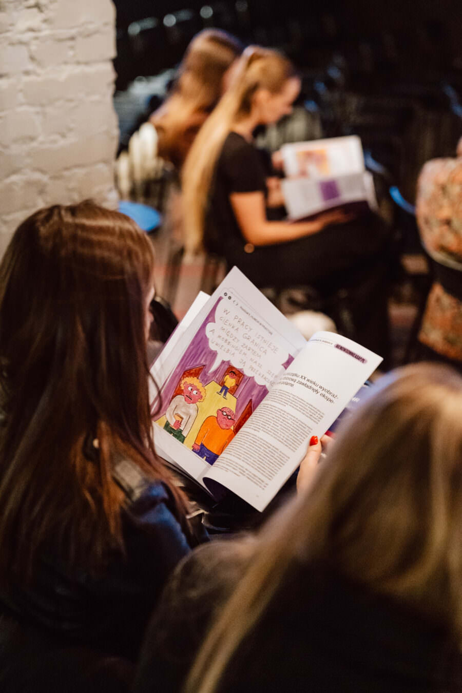 Osoba siedzi w słabo oświetlonym pokoju i czyta czasopismo z kolorowymi ilustracjami, podczas gdy inne osoby w pobliżu również czytają podobne czasopisma. Otoczenie wydaje się swobodne i zrelaksowane.