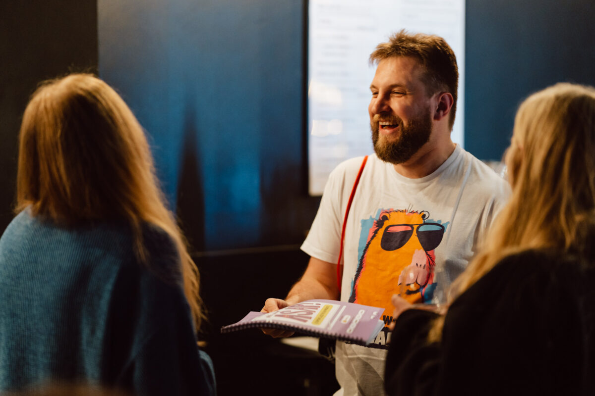 Nie mow do mnie ASAP Klub Komediowy 008 Ein bärtiger Mann, der ein weißes T-Shirt mit bunten Tiermotiven trägt, lächelt und hält ein Notizbuch in der Hand, während er mit zwei Personen in einem schwach beleuchteten Raum spricht.