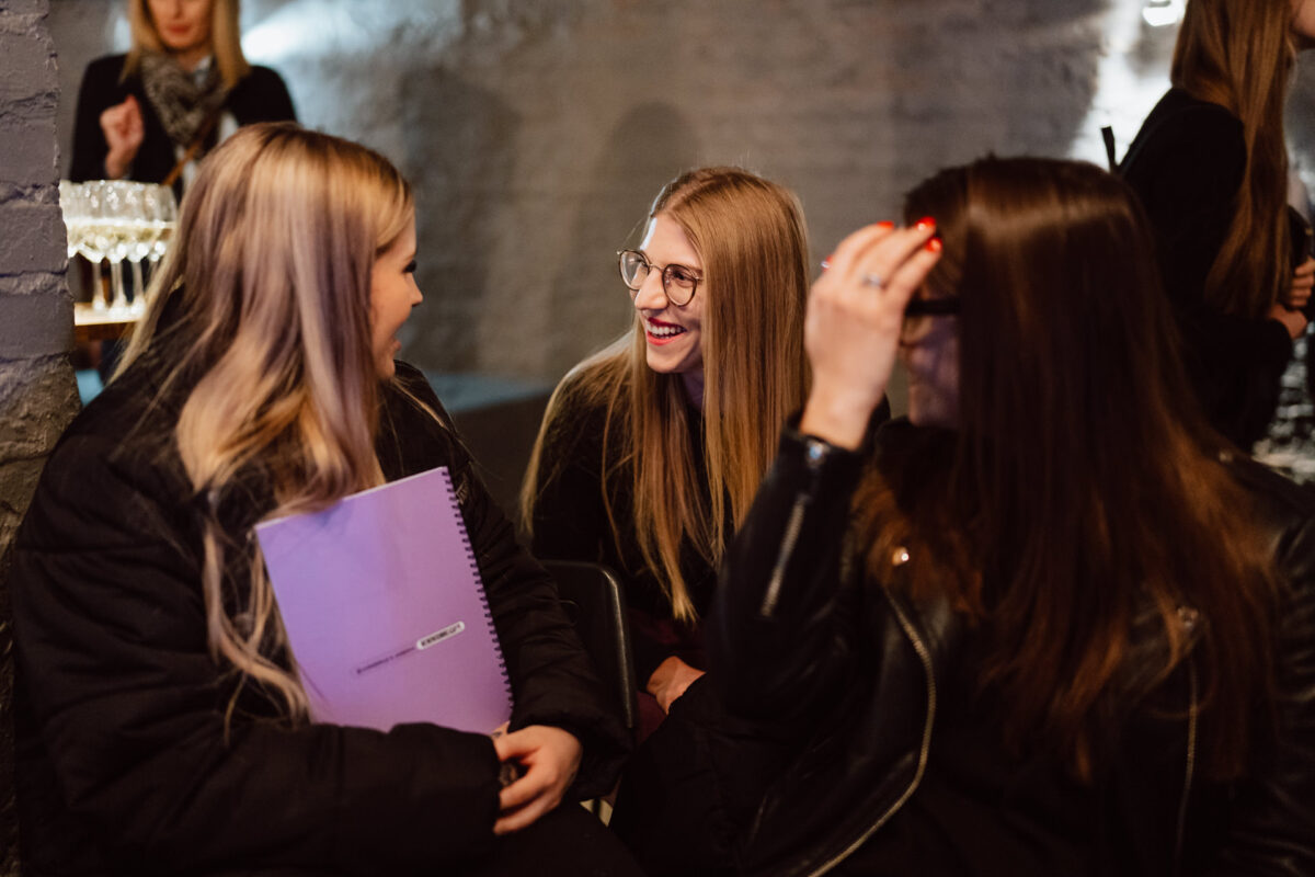 Nie mow do mnie ASAP Klub Komediowy 009 Drei junge Frauen sitzen zusammen in einem Raum, lächeln und unterhalten sich. Eine hält einen lila Notizblock, eine andere trägt eine Brille und die dritte korrigiert ihr Haar. Der Hintergrund ist sanft beleuchtet und die anderen Personen sind unscharf.