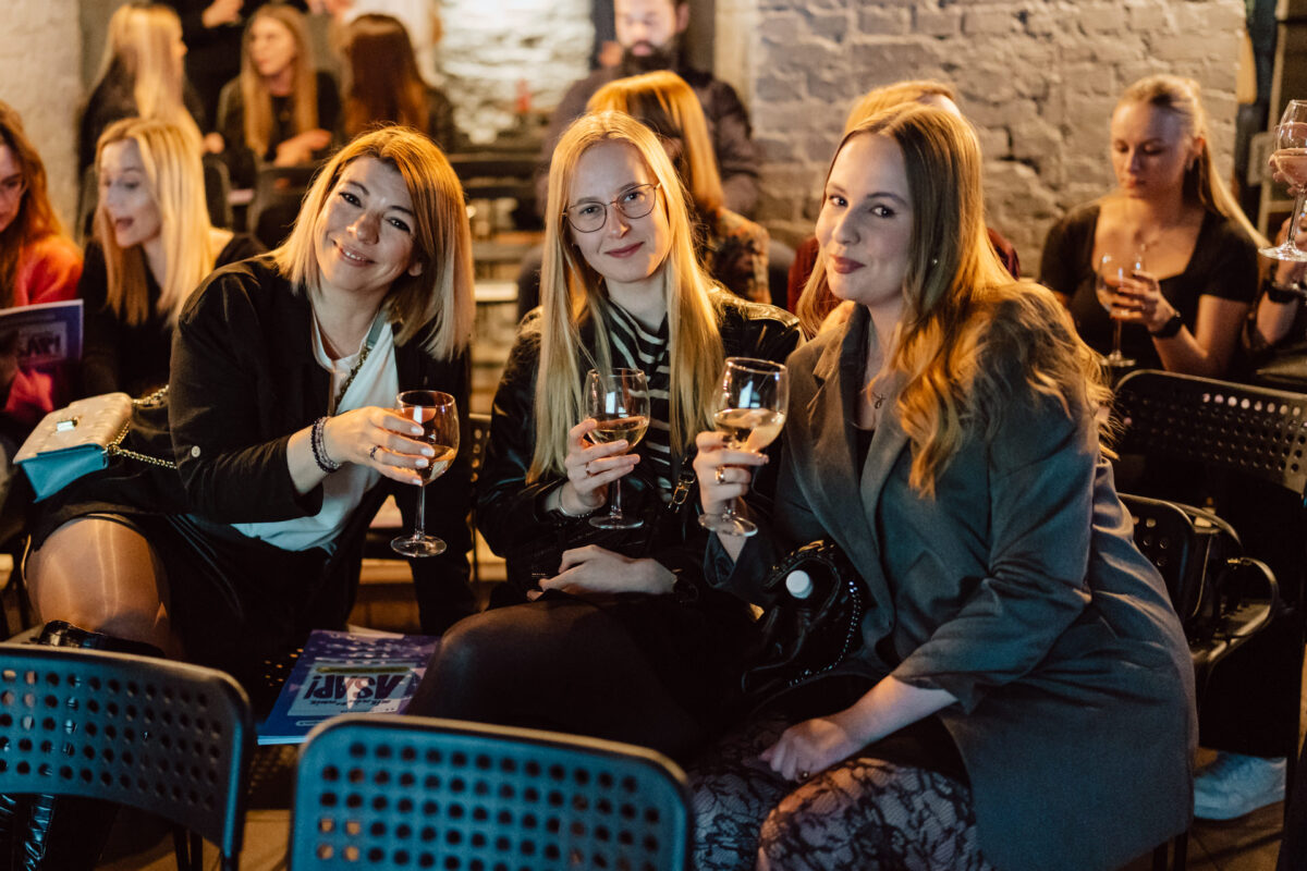 Nie mow do mnie ASAP Klub Komediowy 013 Drei Frauen, die auf einer geschlossenen Party zusammensitzen, lächelnd und mit Weingläsern in der Hand, umgeben von anderen, die in Stuhlreihen in einer warm beleuchteten, ungezwungenen Atmosphäre sitzen.