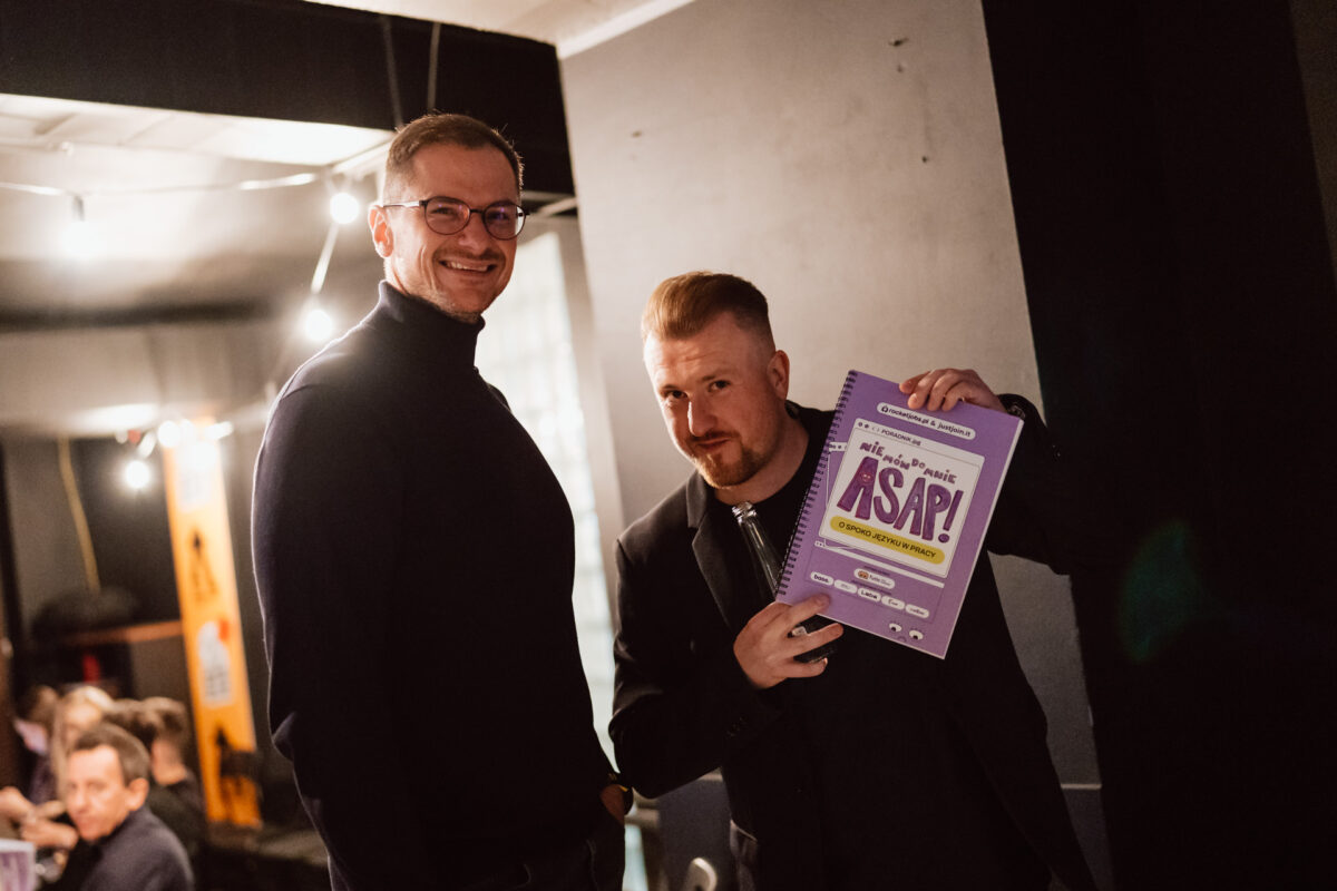 Nie mow do mnie ASAP Klub Komediowy 014 Zwei Männer stehen im Raum, einer lächelt in die Kamera, während der andere ein lila Plakat mit der Aufschrift "ASAP!" hält. Im Hintergrund sitzen Menschen an Tischen.