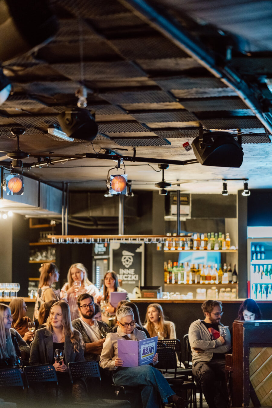 Nie mow do mnie ASAP Klub Komediowy 016 Eine Gruppe von Menschen sitzt und steht in einer gemütlichen Bar, hinter der Regale mit Flaschen stehen. Einige unterhalten sich, andere lesen Programme, und die Atmosphäre ist dank der sanften Beleuchtung warm und entspannt.