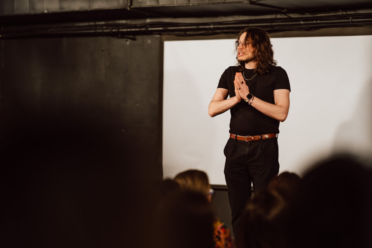 Nie mow do mnie ASAP Klub Komediowy 018 Eine schwarz gekleidete Person mit langen Haaren und einer Brille steht auf der Bühne vor dem Publikum und scheint mit gefalteten Händen zu sprechen oder aufzutreten.