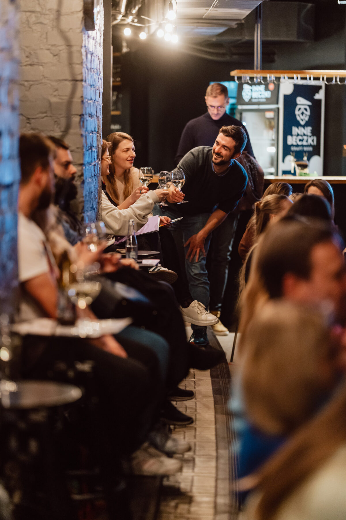 Nie mow do mnie ASAP Klub Komediowy 020 Eine Gruppe von Menschen sitzt dicht beieinander in einer gemütlichen, schwach beleuchteten Bar. Sie lächeln und erheben ihre Gläser zu einem lebhaften Trinkspruch, während andere sich unterhalten und die Atmosphäre genießen.