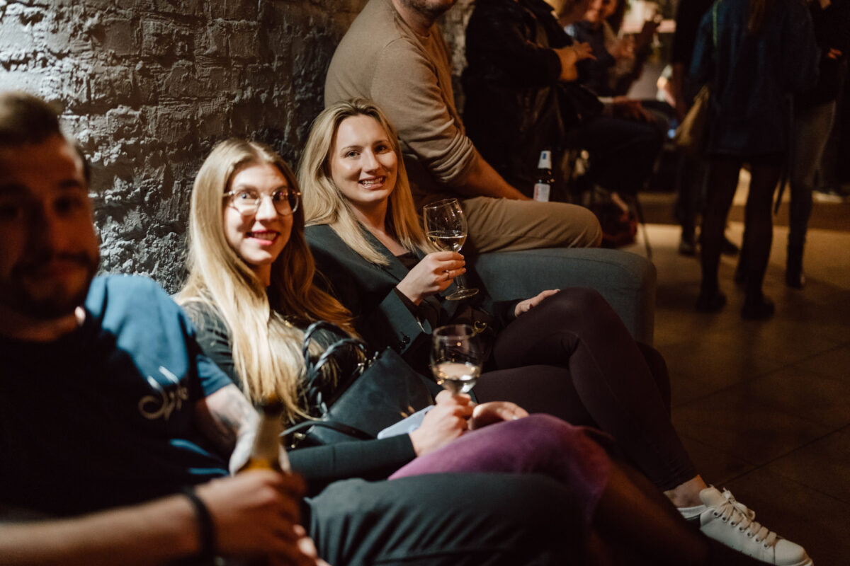 Nie mow do mnie ASAP Klub Komediowy 021 Drei Personen sitzen auf einem Sofa während eines schwach beleuchteten geselligen Beisammenseins. Die beiden Frauen in der Mitte lächeln und halten Weingläser in der Hand, während sich andere Personen im Hintergrund unter sie mischen.