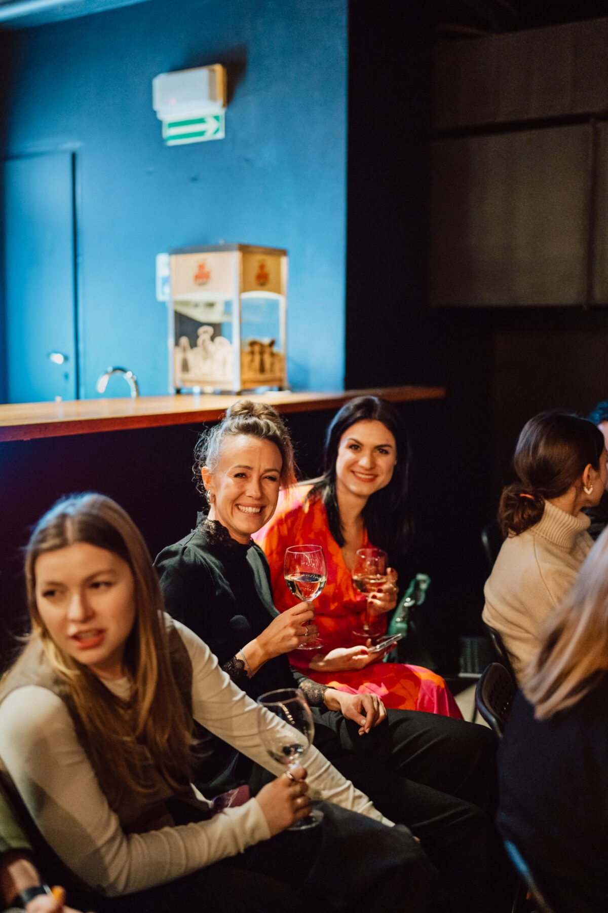 Nie mow do mnie ASAP Klub Komediowy 022 Drei Frauen sitzen in einer Bar oder Lounge, halten Weingläser und lächeln. Der Hintergrund ist schwach beleuchtet, mit einem Holztresen und einer Kiste darauf. Die Atmosphäre scheint entspannt und gesellig zu sein.