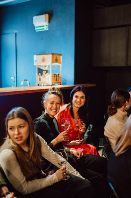 Trzy kobiety siedzą w barze lub salonie, trzymając kieliszki do wina i uśmiechając się. Tło jest słabo oświetlone, z drewnianą ladą i pudełkiem na niej. Atmosfera wydaje się zrelaksowana i towarzyska.