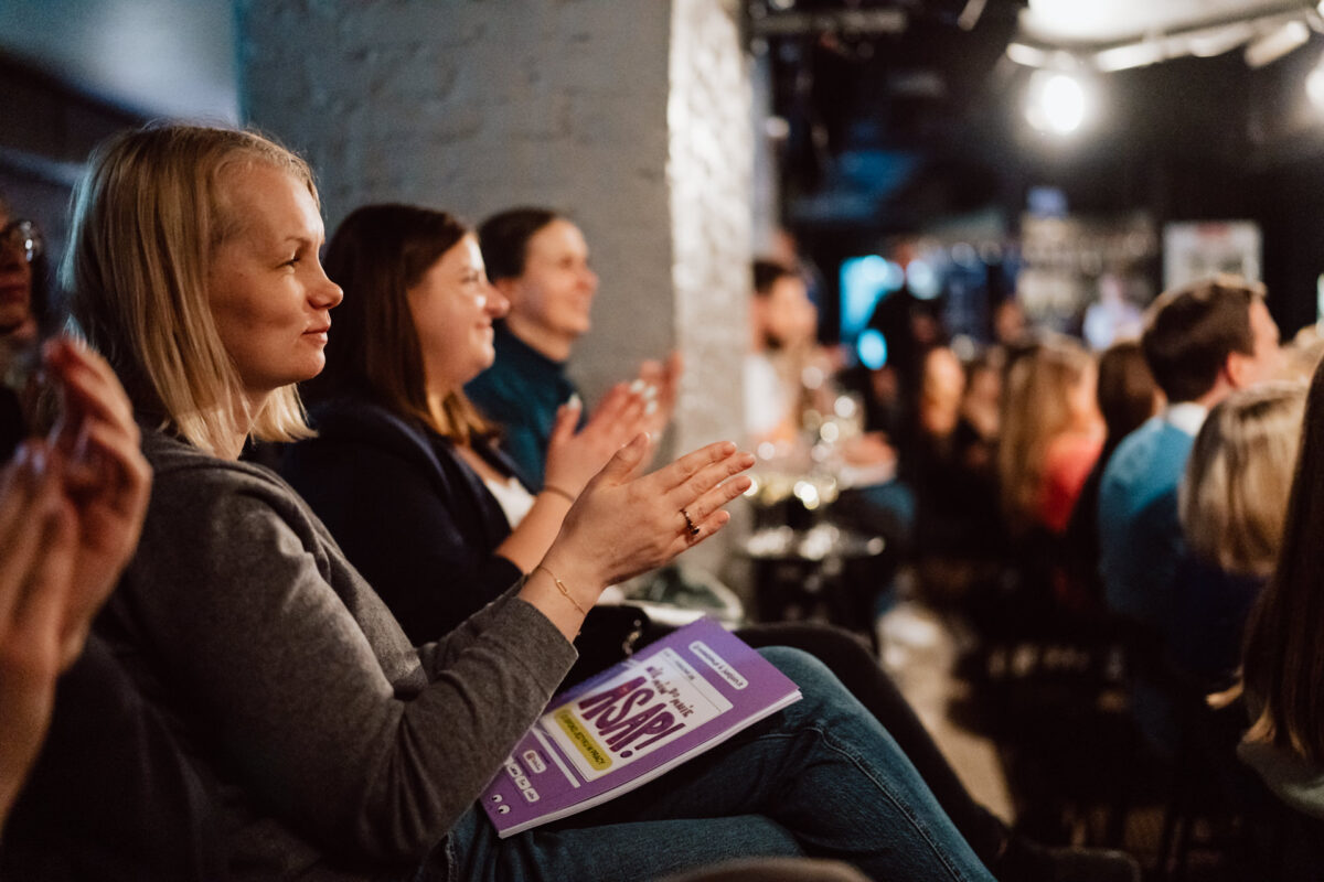 Nie mow do mnie ASAP Klub Komediowy 024 Eine Gruppe von Menschen sitzt in dem schwach beleuchteten Auditorium, schaut aufmerksam zu und klatscht. Eine Frau im Vordergrund hält ein violettes Programmheft mit der Aufschrift 'ASKR' in der Hand. Die Atmosphäre wirkt lebendig und engagiert.
