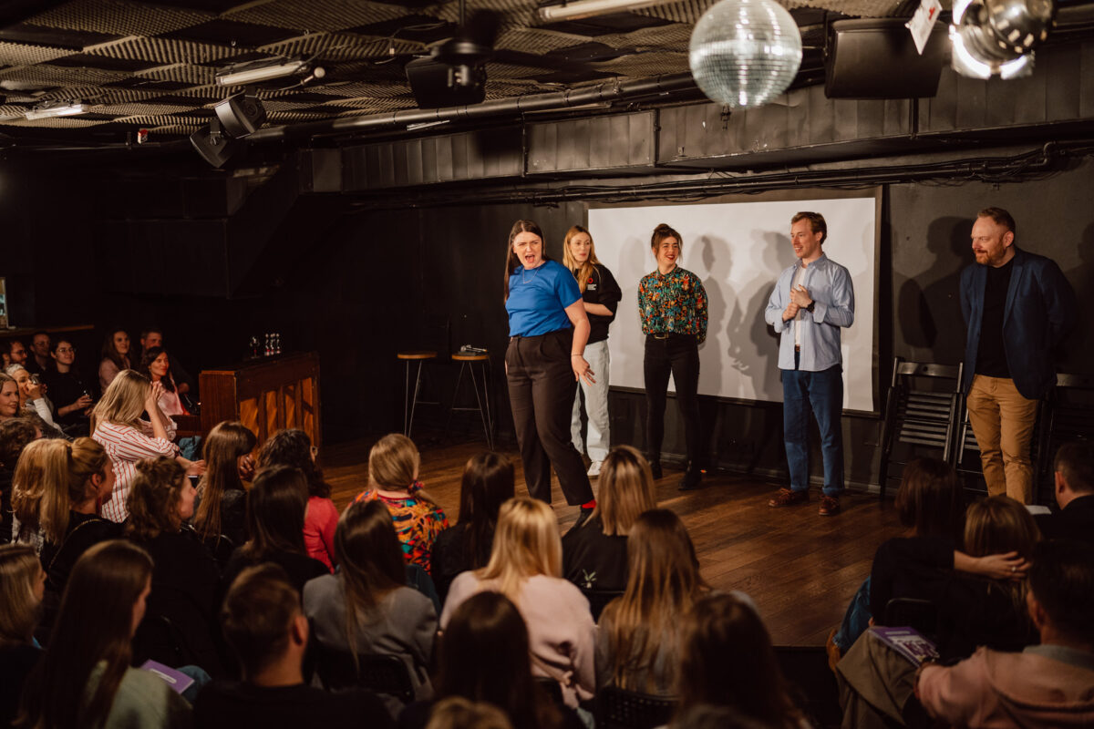 Nie mow do mnie ASAP Klub Komediowy 025 Fünf Personen treten auf der Bühne vor einem Publikum auf. Eine Person steht selbstbewusst an der Spitze, während die anderen lächeln und zusehen. Es scheint sich um Comedy oder Improvisation in einem kleinen, intimen Rahmen zu handeln.