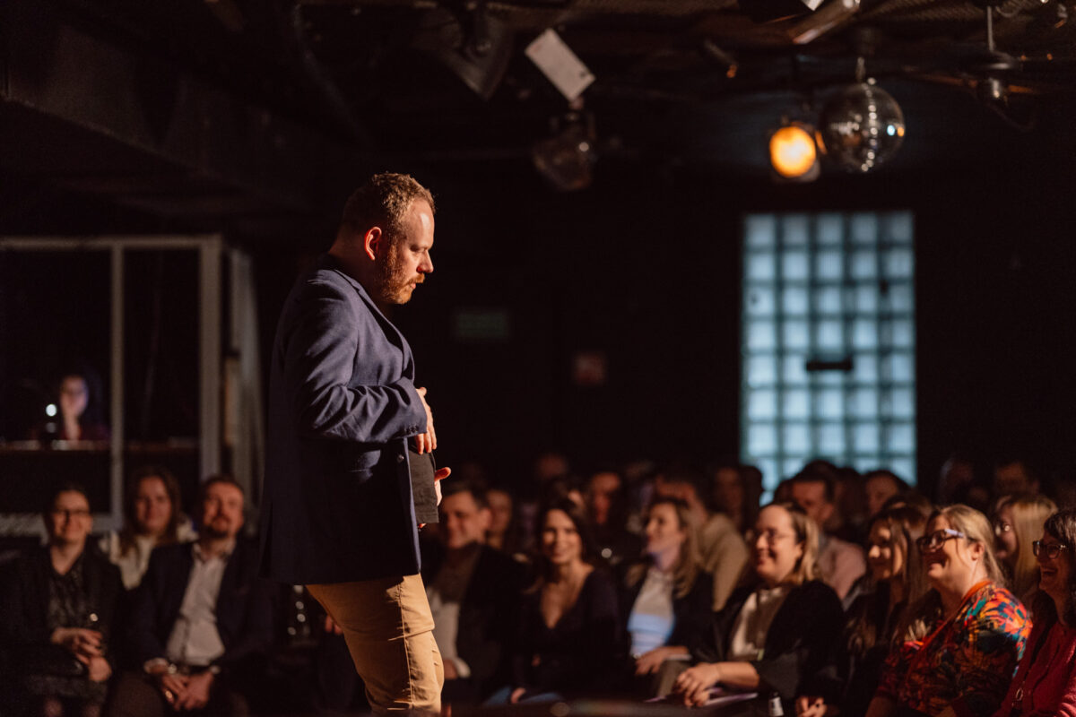 Nie mow do mnie ASAP Klub Komediowy 028 Ein Mann in einer blauen Jacke tritt auf der Bühne vor einem aufmerksam sitzenden Publikum in einem schwach beleuchteten Saal auf. Im Hintergrund sind eine Discokugel, Bühnenscheinwerfer und eine Person an einem Schaltpult zu sehen.