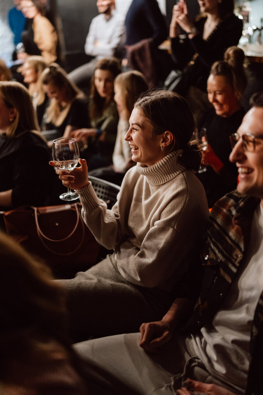 Nie mow do mnie ASAP Klub Komediowy 032 Eine Frau in einem hellen Pullover lächelt und hält ein Glas Wein in der Hand, während sie inmitten einer Gruppe von Menschen bei einem gesellschaftlichen Ereignis sitzt. Andere um sie herum lächeln ebenfalls und sind in den Moment vertieft.
