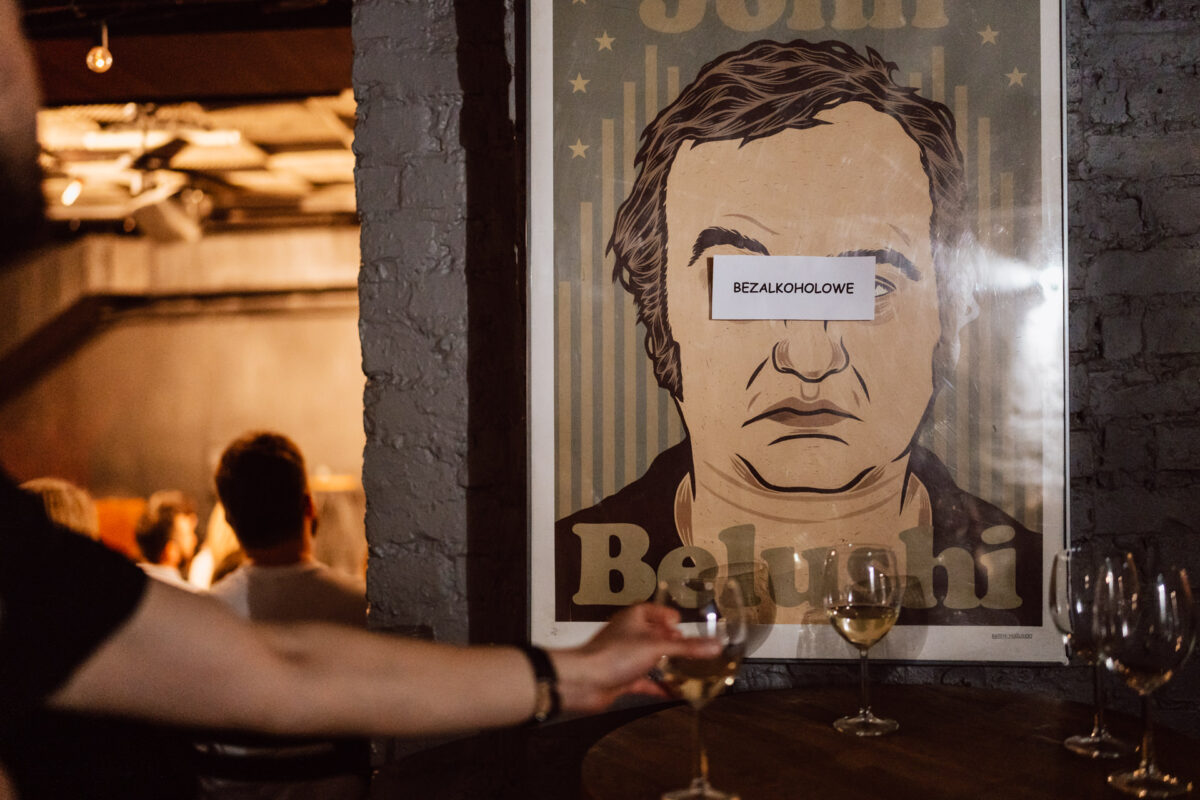 Nie mow do mnie ASAP Klub Komediowy 034 Eine Person greift nach einem Glas Wein auf einem Tisch in einem schwach beleuchteten Raum. Im Hintergrund ist das Poster eines Mannes zu sehen, über dessen Augen das Wort 'OHNE Alkohol' steht, und auf dem Tisch stehen mehrere leere Weingläser.
