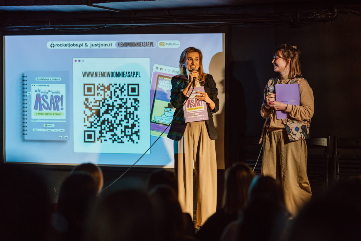 Nie mow do mnie ASAP Klub Komediowy 035 Zwei Frauen stehen auf der Bühne vor einem Bildschirm, der einen QR-Code, einen Link zu einer Website und eine Werbegrafik zeigt. Eine von ihnen hält ein Mikrofon und Papier in der Hand, die andere lächelt und hält einen Ordner. Die Zuschauer sind im Vordergrund zu sehen.
