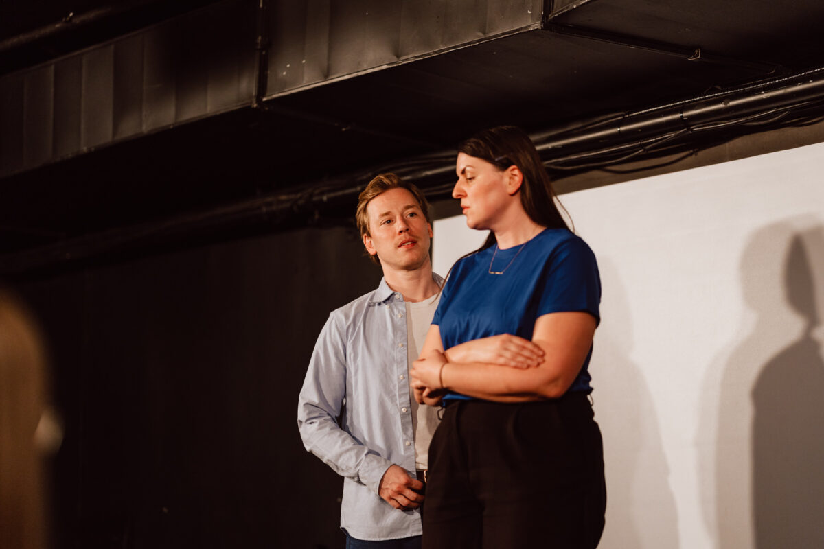 Nie mow do mnie ASAP Klub Komediowy 043 Ein Mann in einem hellen Hemd steht hinter einer Frau in einem blauen Hemd mit verschränkten Armen. Sie scheinen auf einer Bühne zu stehen, mit einem schlichten weißen Hintergrund und sichtbaren Rohren über ihnen.