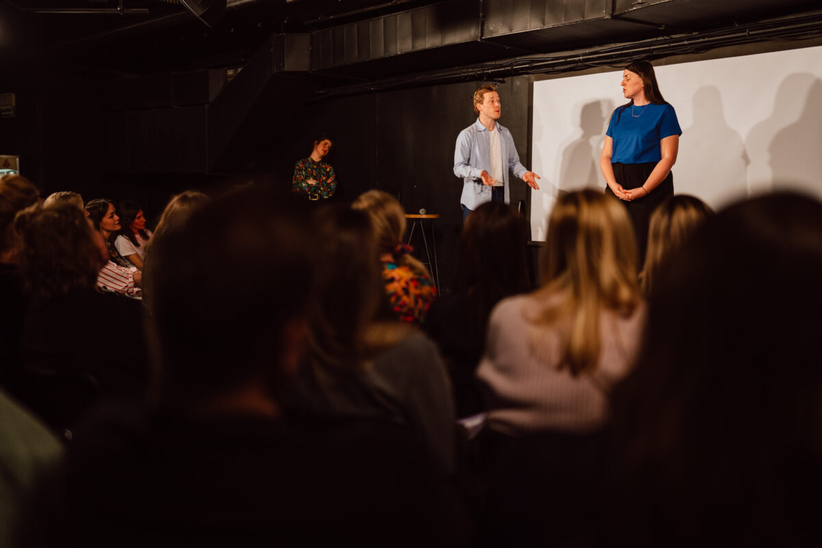 Nie mow do mnie ASAP Klub Komediowy 044 Ein Mann und eine Frau treten auf der Bühne vor einem sitzenden Publikum in einem schwach beleuchteten Raum auf. Eine weitere Person steht im Hintergrund, teilweise sichtbar. Das Publikum schaut gebannt zu.