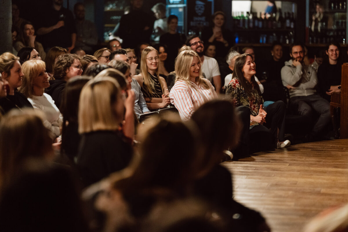 Nie mow do mnie ASAP Klub Komediowy 048 Eine Gruppe von Menschen sitzt dicht beieinander in einem schwach beleuchteten Raum, lächelt und lacht, während sie eine Live-Performance oder einen Redner verfolgen, wodurch eine lebhafte und engagierte Atmosphäre entsteht.